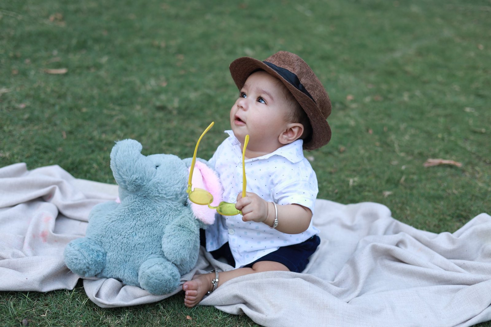 Six month old in a brown hat sitting on a picnic blanket with a toy elephant