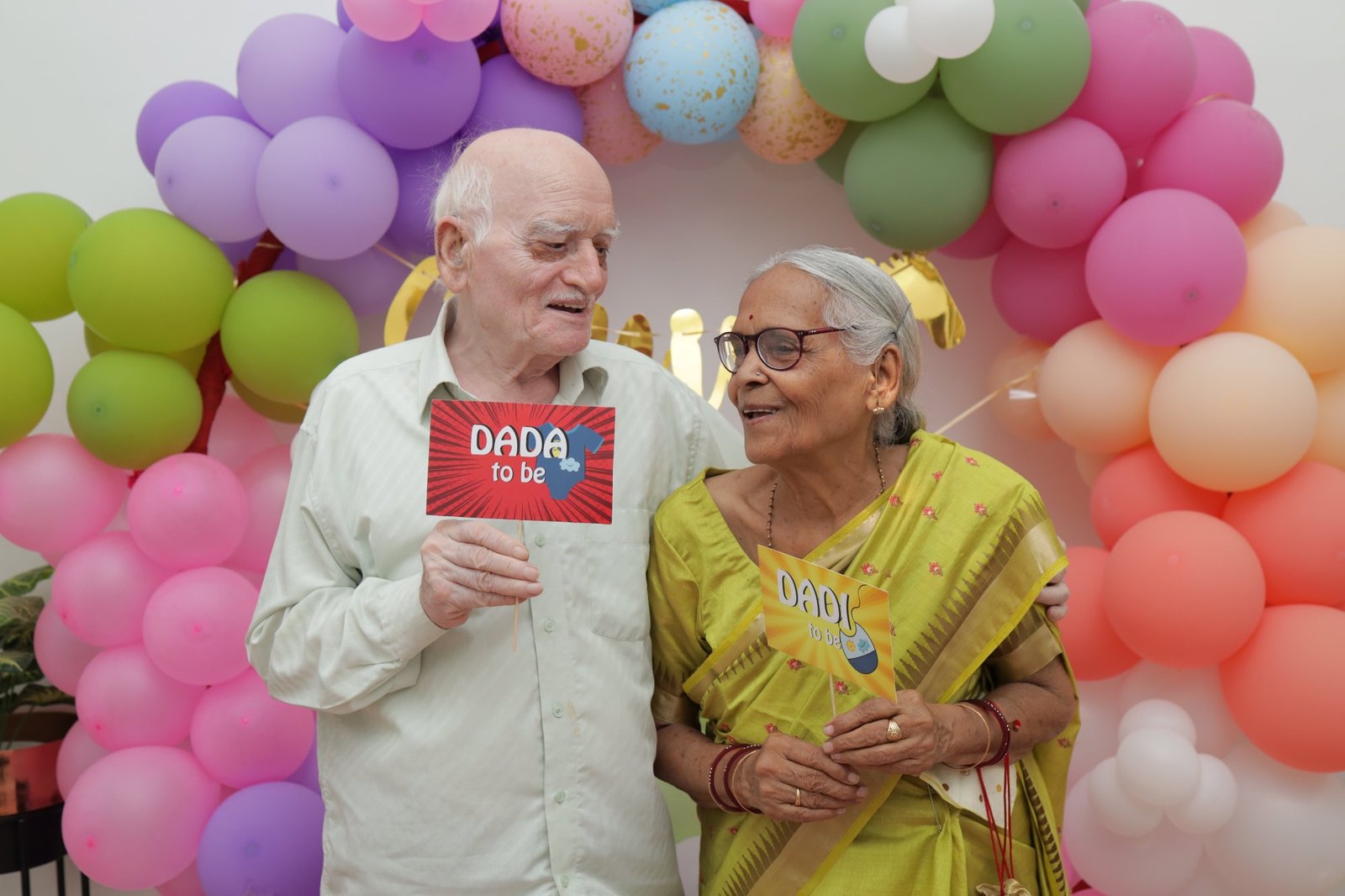 Grandparents-to-be holding a celebration sign in front of the balloon wall