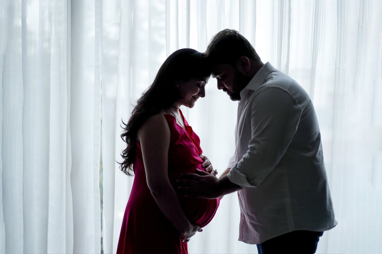 Expectant couple leaning their foreheads together in soft window light