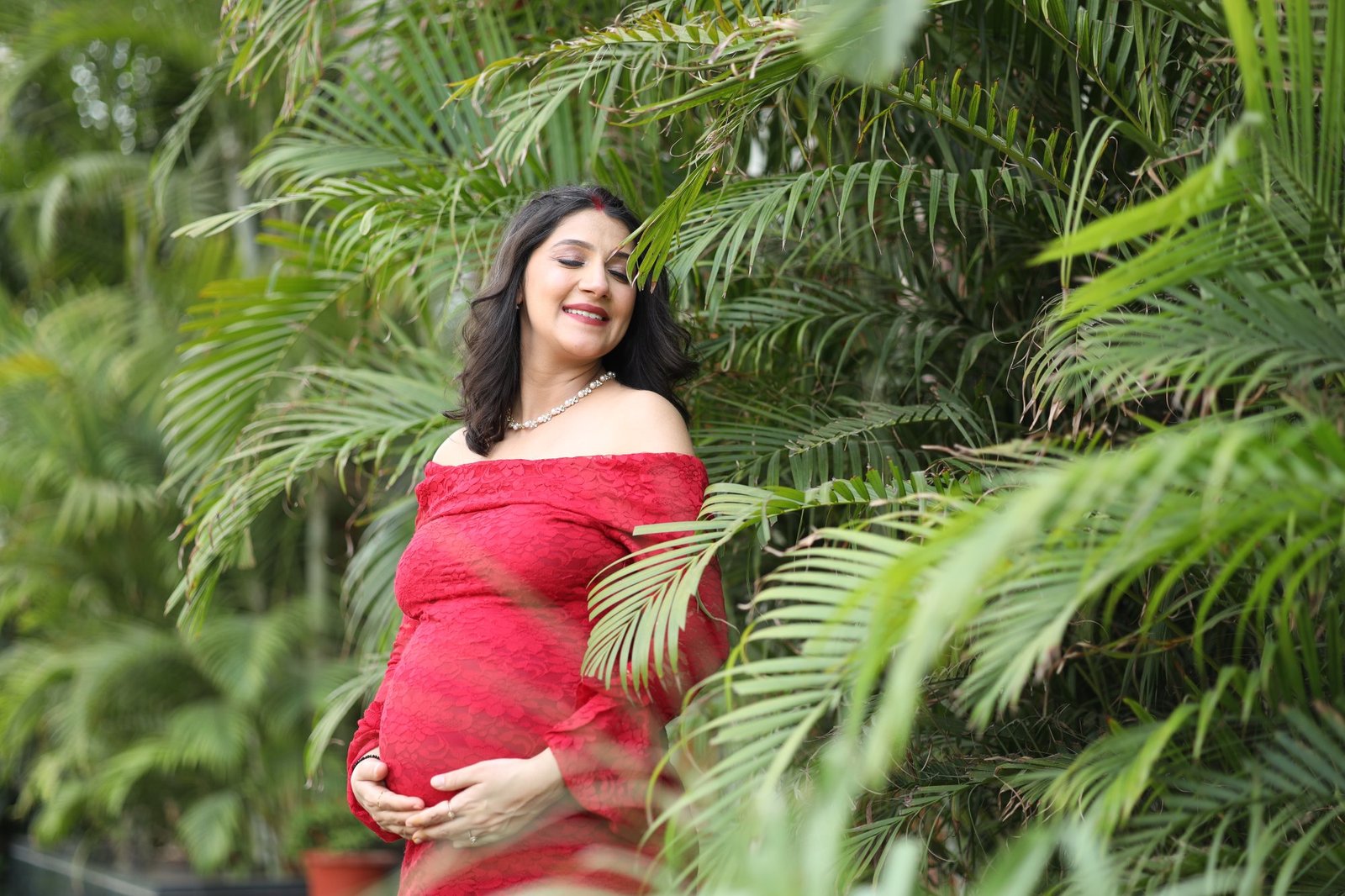 Expectant mother in a red gown cradling her bump among green ferns