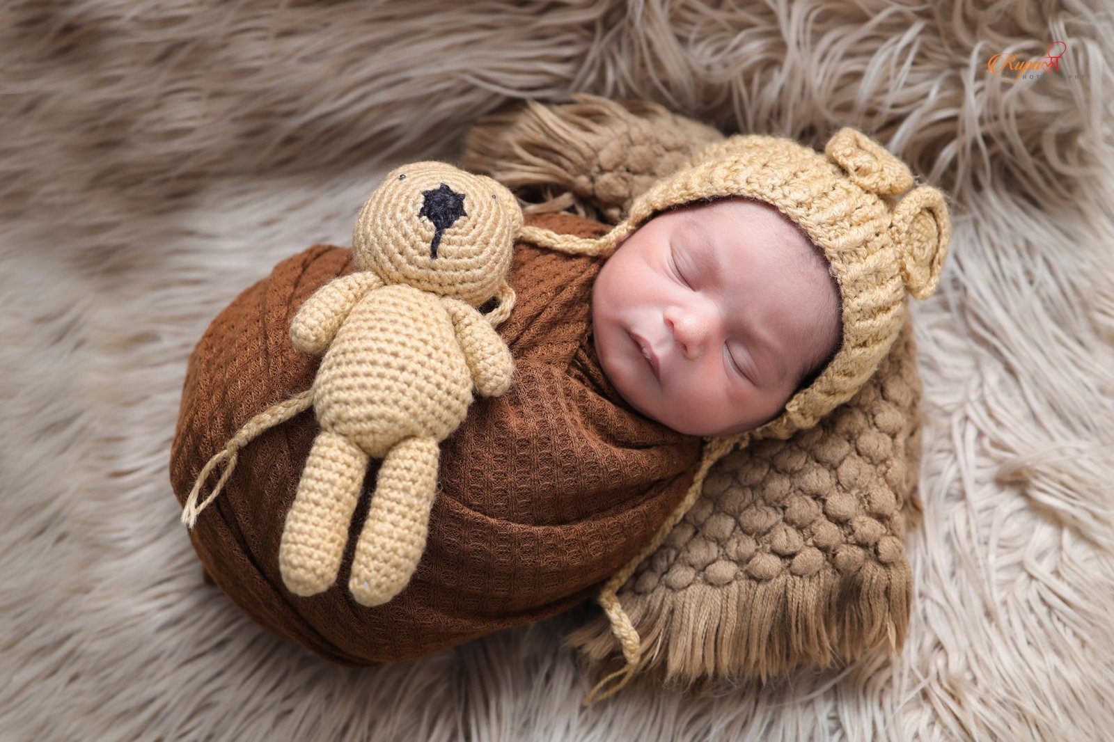 Newborn in a knitted bonnet asleep on fur beside a small teddy bear