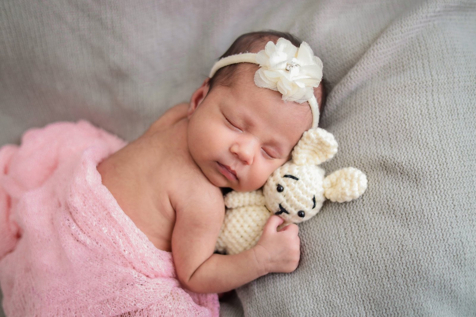 Newborn in a pink wrap and headband holding a small knitted bear
