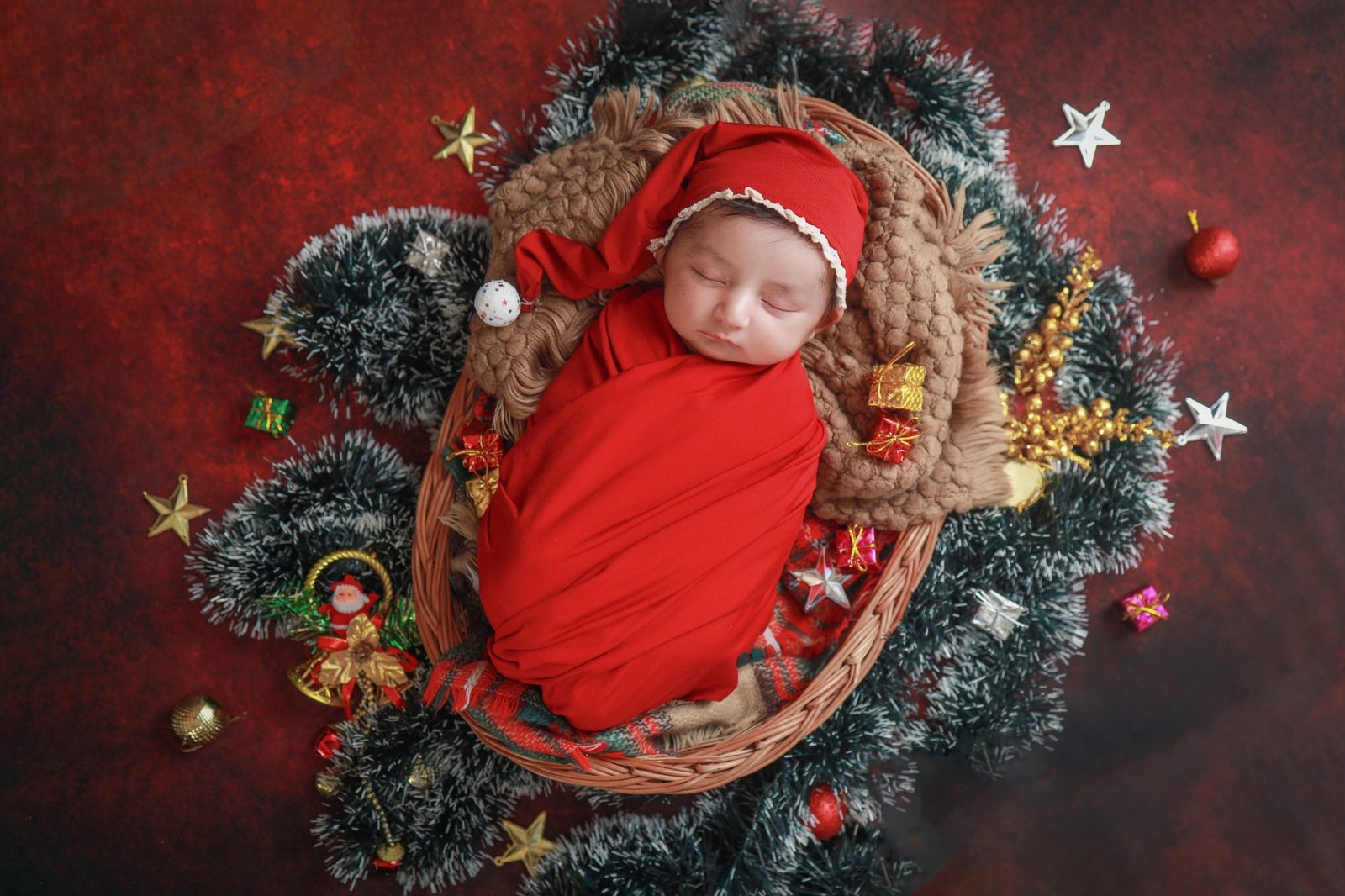 Newborn in a red wrap and knitted hat asleep inside a festive wreath