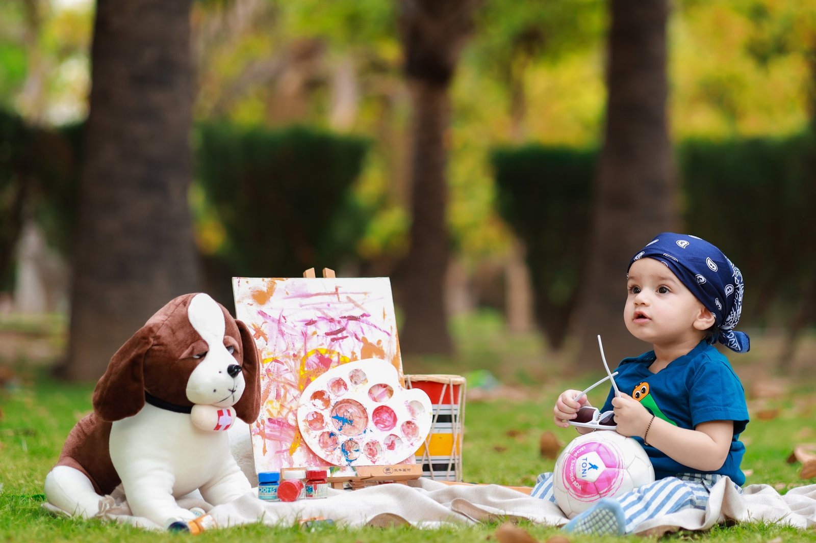 Toddler in a bandana on a picnic rug with a large toy dog and paints