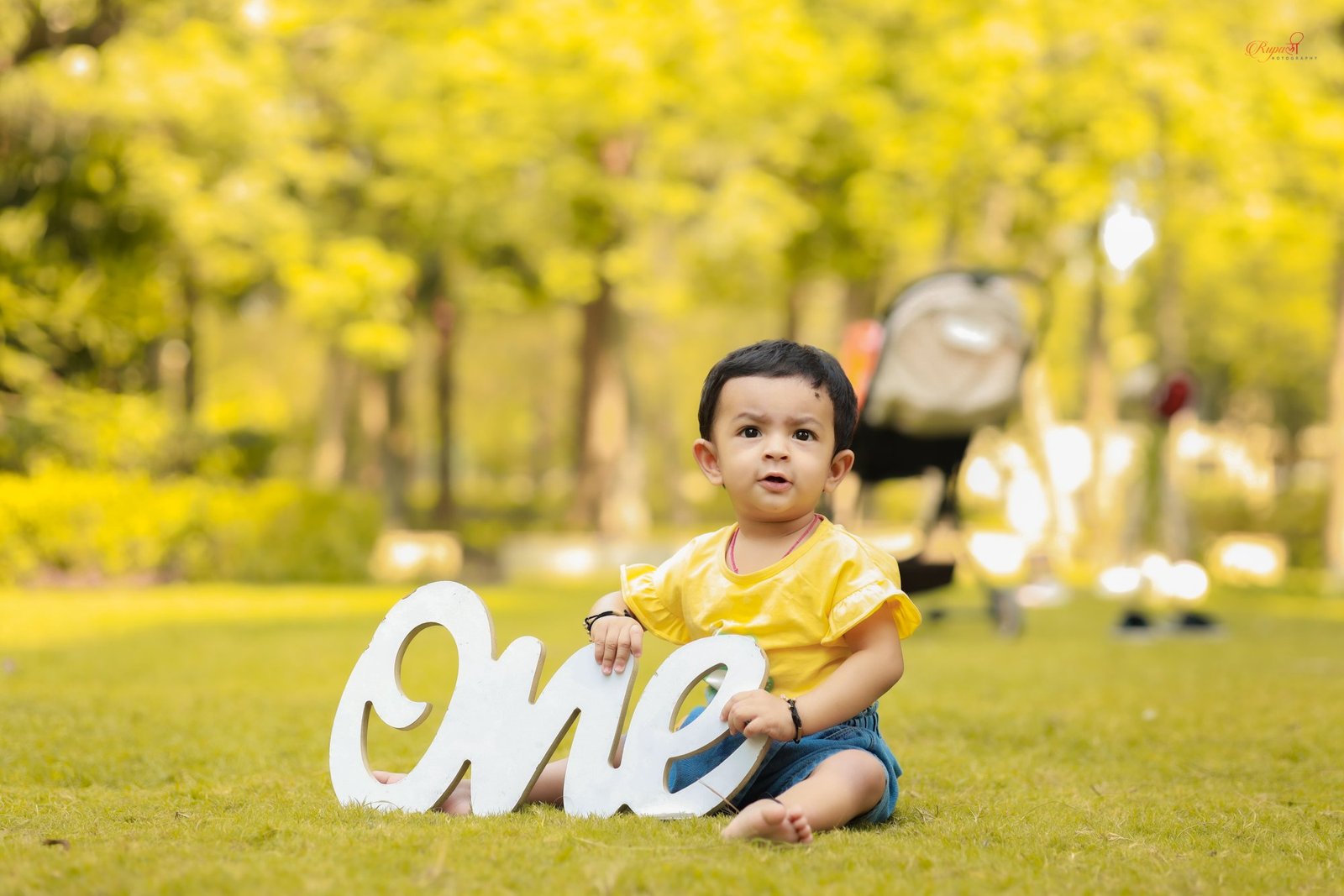 Toddler in a yellow top seated on the grass beside a ONE sign