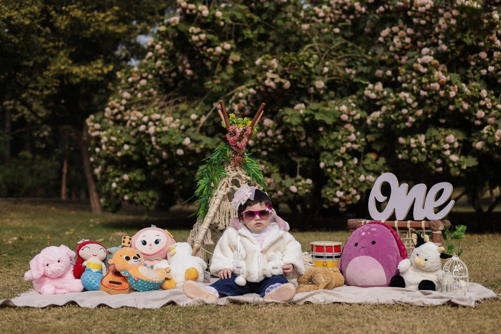 Toddler seated on a picnic rug at an outdoor teepee surrounded by soft toys
