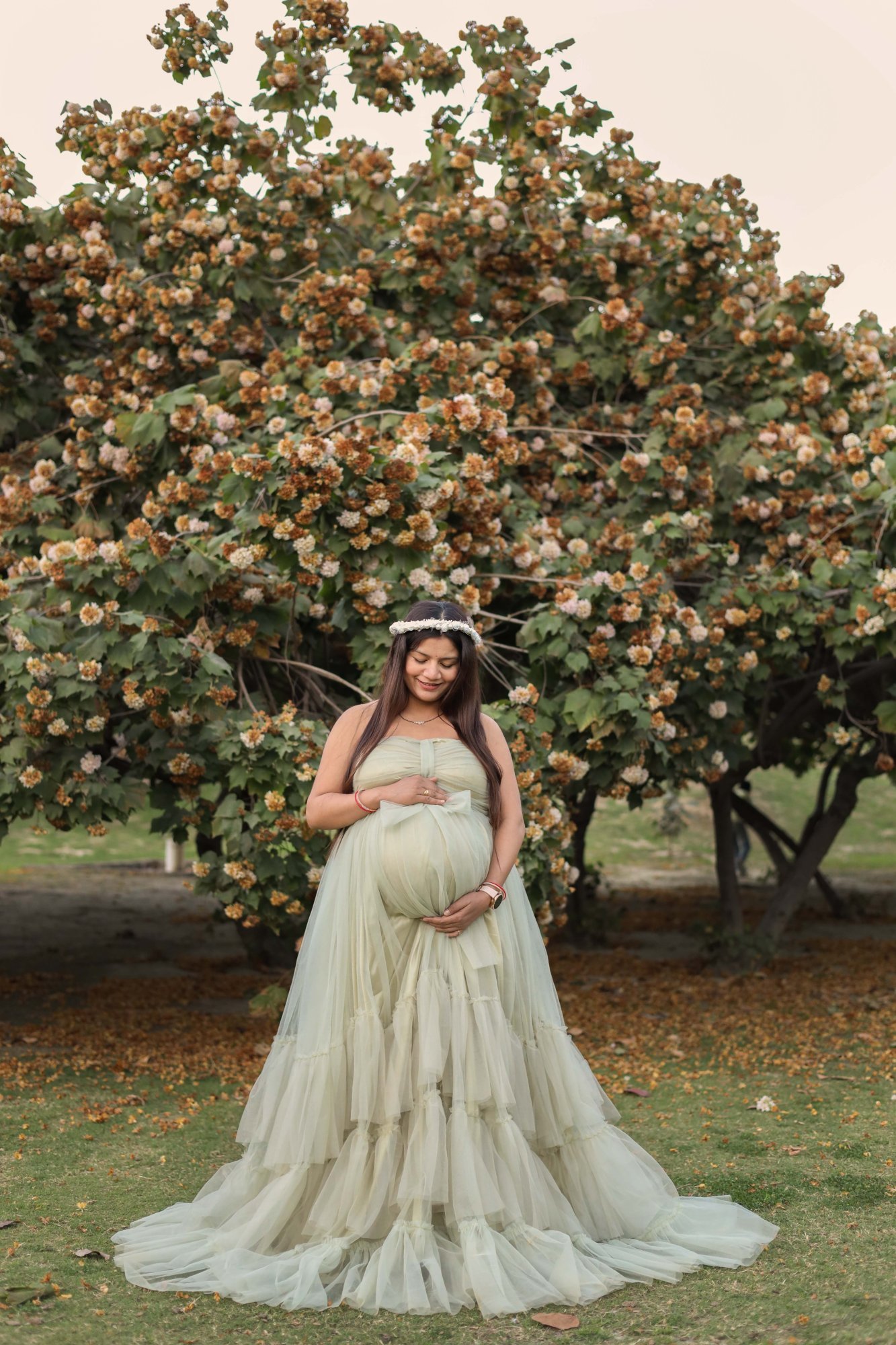 Expectant mother in a flowing cream gown beside a blossoming tree